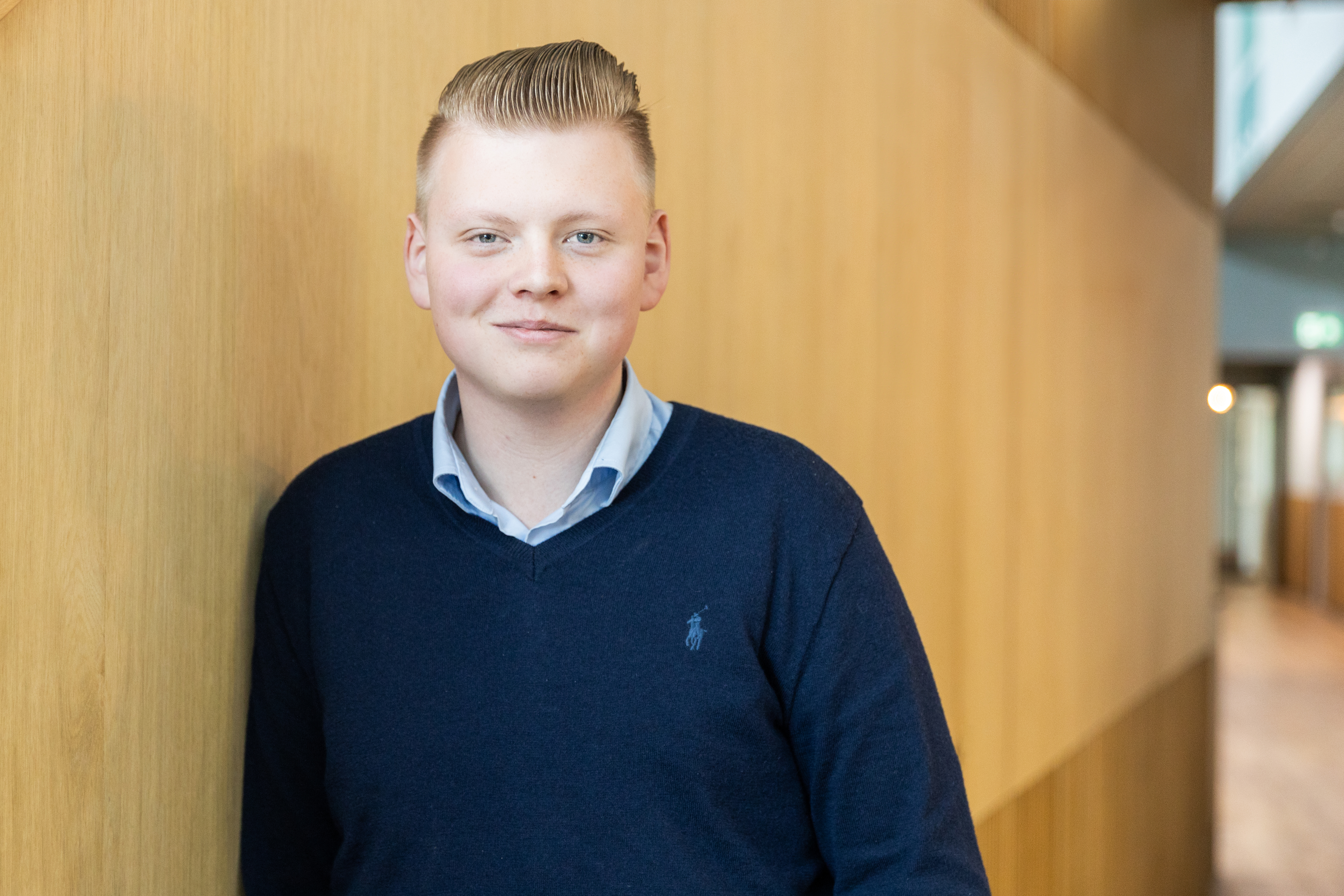 Student staat ontspannen tegen een houten wand in een onderwijsomgeving, passend bij een Erasmus-uitwisseling.