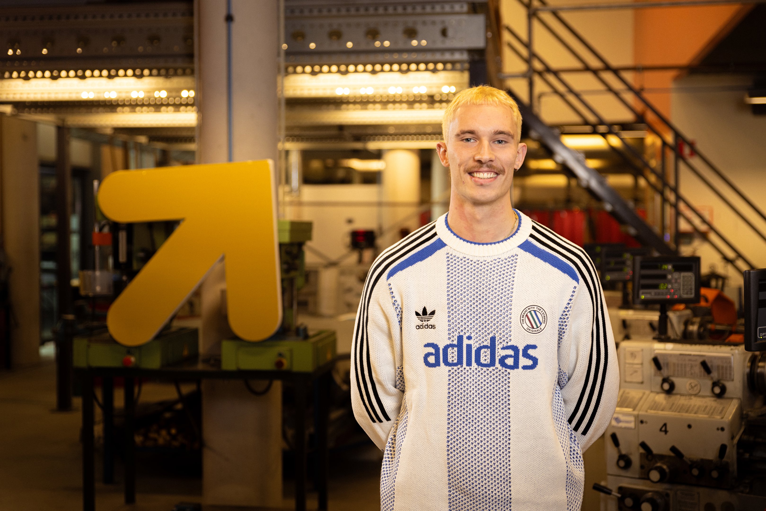 Student poseert in technische leeromgeving met machines en een opvallend gele pijl op de achtergrond