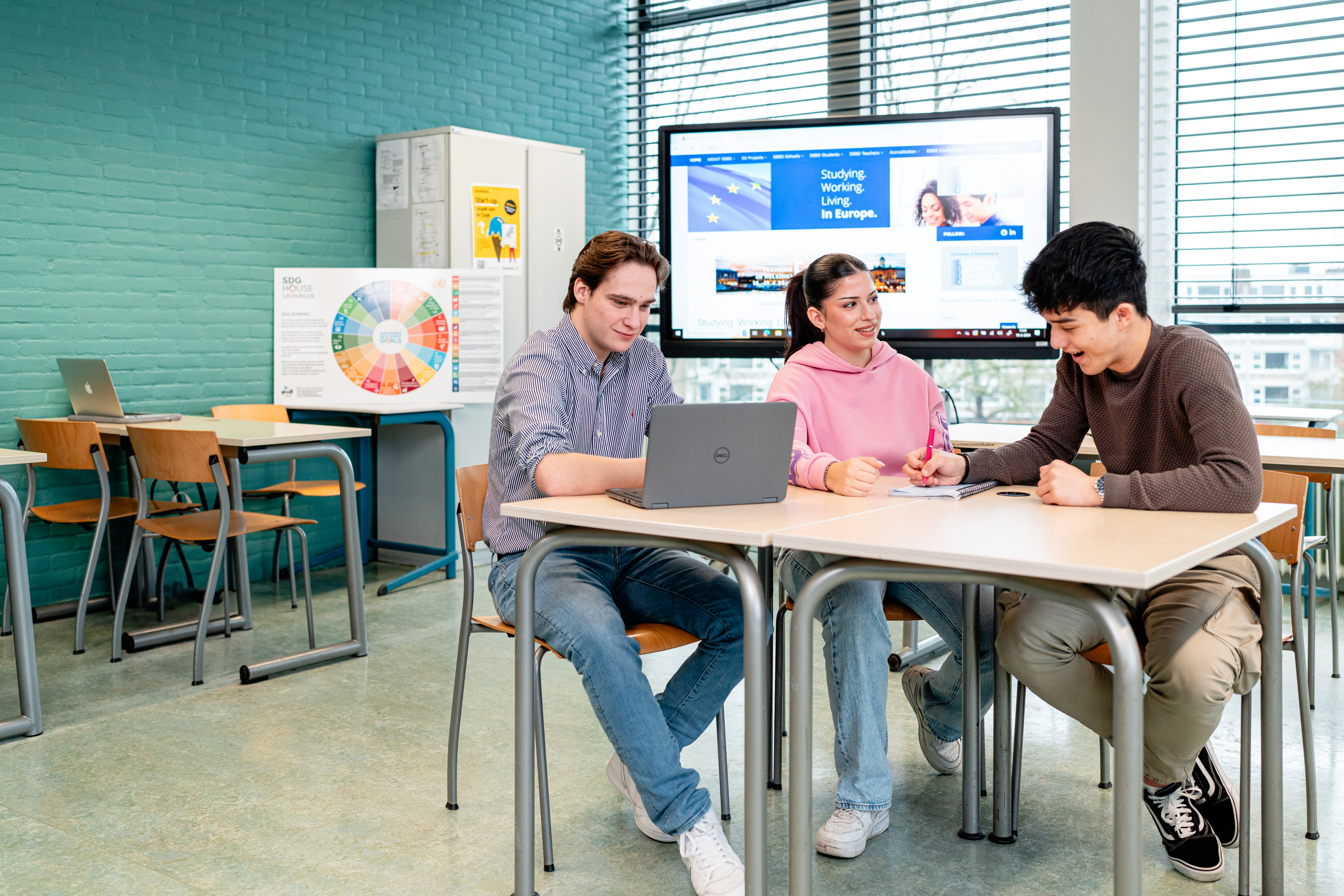 Studenten werken samen aan een opdracht over internationale handel in een lokaal met scherm en laptop